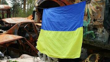 The national flag of Ukraine against the background of ruins and rusty metal of destroyed cars. War in Ukraine. A blue-yellow flag waving in the wind under the sun in a bomb-ravaged city. video