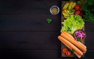 Ingredients for hot dog with  sausage, pickled cucumber, tomatoes, red onions and lettuce on dark wooden background. Summer hotdog. Top view photo