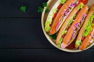 Hot dog with  sausage, pickled cucumber, tomatoes, red onions and lettuce on dark wooden background. Summer hotdog. Top view photo