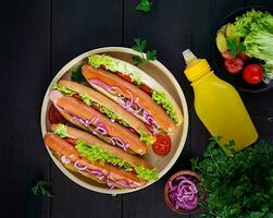 Hot dog with  sausage, pickled cucumber, tomatoes, red onions and lettuce on dark wooden background. Summer hotdog. Top view photo