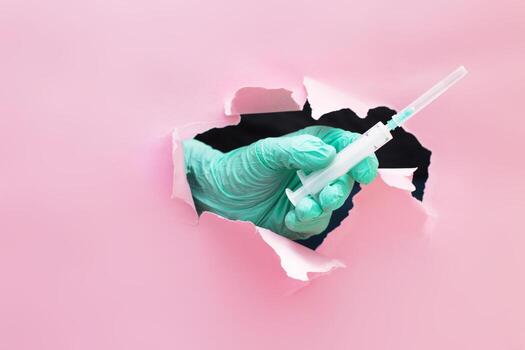 doctor's hand holds out a syringe with a vaccine through a hole in torn paper on a pink background photo