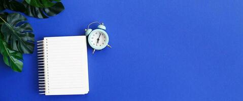 Top view of blank note paper, alarm clock and monstera leaf on blue background photo