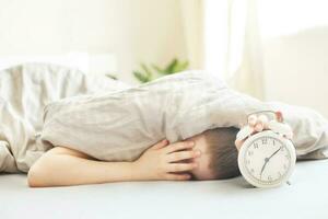 boy lying on the bed and stopping alarm clock in morning. childs hand reaching for the alarm clock to turn it off. photo
