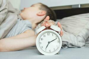 boy lying on the bed and stopping alarm clock in morning. childs hand reaching for the alarm clock to turn it off. photo