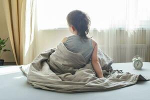 little boy sitting in bed at home in the morning on a window background with alarm clock photo