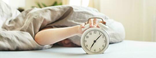 boy lying on the bed and stopping alarm clock in morning. childs hand reaching for the alarm clock to turn it off. photo