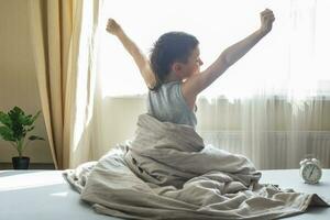 little boy sitting and stretching in bed at home in the morning on a window background with alarm clock photo