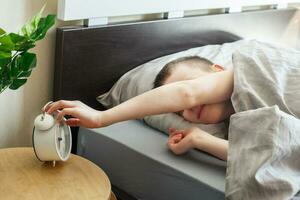 boy lying on the bed and stopping alarm clock in morning. childs hand reaching for the alarm clock to turn it off. photo