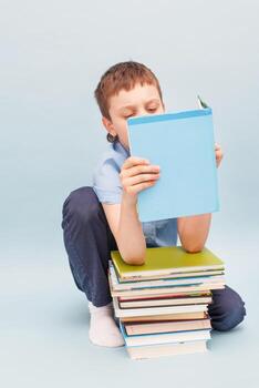Schoolboy is sitting with a stack of books and reading isolated on blue background photo