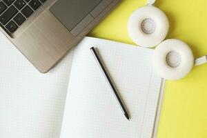 Top view open notepad, headphones pen and laptop on yellow background. workplace photo