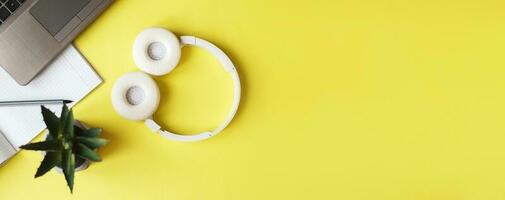 Top view open notepad, headphones pen and laptop on yellow background. workplace. banner photo