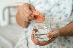 Asian senior woman patient holding and washing denture in water cleanser glass for good chewing. photo