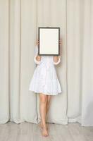 woman in dress holding blank frame, beige curtain background photo