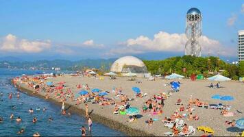 Panoramic view from pier to Batumi coast with people relax swim in beach and tourist attractions with black sea background. Vacation holidays in caucasus. Georgian summer holiday video