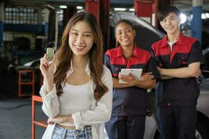 Young Asian female customer shows car key with automotive mechanic workers, happy smile in quality trust at maintenance garage, professional vehicle service center, fix check and repair auto industry. photo