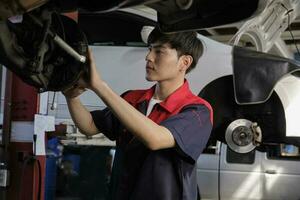 One young expert Asian male automotive mechanic technician is screwing car wheel nuts on lifting with wrench for repair at garage. Vehicle maintenance service works, industry occupation business jobs. photo