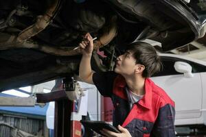 profesional joven asiático masculino motor mecánico inspecciona tren de aterrizaje de eléctrico coche levantado por máquina elevadora Jack para reparar a cochera, automotor mantenimiento Servicio trabajos industria ocupación negocio. foto