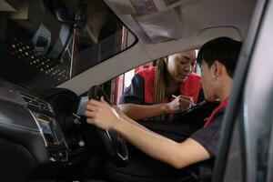 Young male Asian automotive mechanic technician and partner checking maintenance list with tablet in car interior at garage. Vehicle service fix and repair works, industrial occupation business jobs. photo