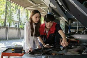 Male automotive mechanical worker explains checklists to Asian customer, repair an EV car, and hybrid engine at maintenance garage, expert electric vehicle service, and occupation auto fix industry. photo