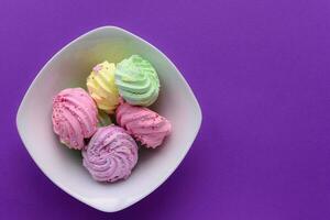 Colorful delicious meringues in white bowl on purple background photo