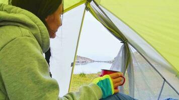 femme réfléchie tenir une tasse de camping et boire dans une tente verte à l'extérieur dans la nature avec vue panoramique sur le lac alpin en automne video