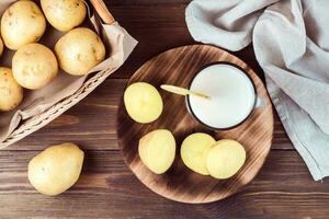 Alternative vegetable vegan potato milk in a glass and potato tubers side by side on a wooden table. Vegetarian food. Top view photo