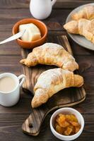 Fresh crispy croissants on a wooden board, a cup of coffee and jam in a bowl on the table. Brunch and coffee break. Vertical view photo