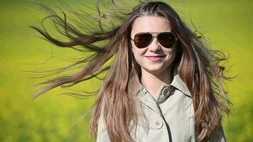 souriant Jeune fille dans des lunettes de soleil avec cheveux soufflant dans vent permanent dans flou la nature Contexte. video