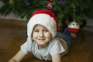 A photo of a beautiful boy in a gray T-shirt and a Santa Claus hat at the Christmas tree, looking into the camera. Portrait in a bright room. Natural, not staged photography.
