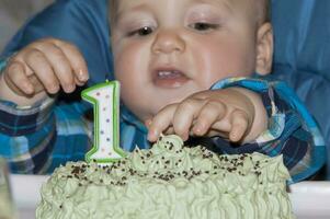 un hermosa bebé celebra su primero cumpleaños y come un pastel con su mano. pastel con el número uno en el mesa en frente de el niño. foto