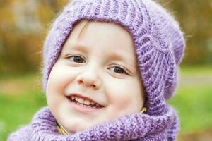 A child in a purple knitted hat and scarf. Portrait of a child against the background of autumn trees. Cute smiling boy. photo