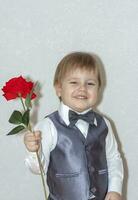 A little boy holds and hands over a red rose, the concept of the Valentine's Day theme. Portrait of a cute boy in a suit with a bow tie. photo