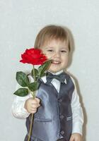 A little boy holds and hands over a red rose, the concept of the Valentine's Day theme. Portrait of a cute boy in a suit with a bow tie. photo