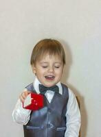 A little boy holds and hands over a red box, a Valentine's Day theme concept. Portrait of a cute boy in a suit with a bow tie. photo