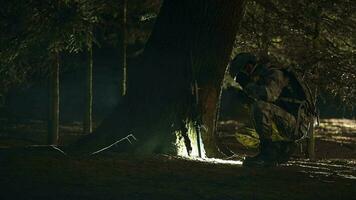noche escena de masculino soldado vistiendo lleno uniforme y engranaje agachado y descansando por árbol mirando alrededor y ajustando casco y gafas de protección, video
