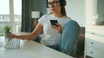 casualmente vestito donna con cuffie Lavorando con un' computer e smartphone a casa nel un' accogliente ambiente. a distanza opera video