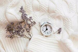 Alarm clock and dry flowers on a cozy white sweater. Wellness time concept. Top view photo