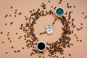 Two cups of black coffee in a circle from coffee beans and a white alarm clock on a beige background. Time to drink coffee concept. Top view photo