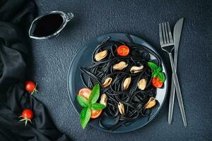 Ready-to-eat black spaghetti with mussels, tomatoes and basil on a plate on a black background. Food photography in dark colors. Top view photo