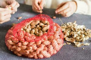 Mesh bag with inshell peanuts and two girls in the background are peeling nuts at the table. Lifestyle photo