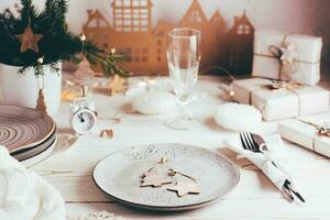 organización de un festivo Navidad mesa. plato con de madera decoraciones, cuchillería, champán vaso y cartulina casa. paisajes de mesa en blanco de madera mesa foto