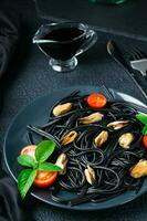 Ready to eat black spaghetti with mussels, tomatoes and basil on a plate and soy sauce on a black background. Food photography in dark colors. Close-up. Vertical view photo