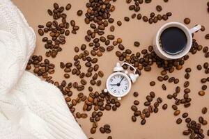 White alarm clock, a cup of black coffee, a cozy sweater and coffee beans on a beige background. Take a break from work for rest. Top view photo