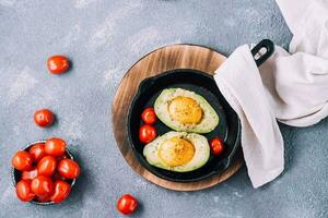 Raw avocado halves, ready to bake, stuffed with egg and cherry tomatoes in a pan on the table. Healthy eating. Flexitarian diet. Top view. Copy space photo