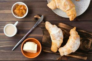 Fresh crispy croissants on a wooden board, a cup of coffee, butter and jam in bowls on the table. Brunch and coffee break. Top view photo