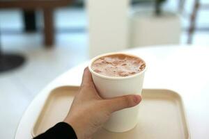 cacao Leche papel taza participación en mujer mano con blanco mesa en café foto