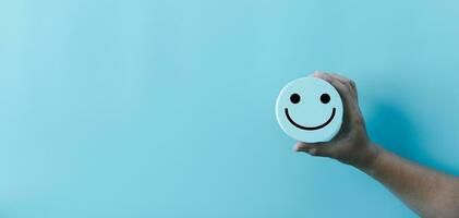 Woman hand holding happy face smile face icon on round blue object. Customer experience and service with satisfaction concept. positive thinking, mental health assessment, world mental health day. photo