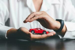 Business woman's hand protecting red toy car on desk. Planning to manage transportation finance costs. Concept of car insurance business, saving buy - sale with tax and loan for new car. photo