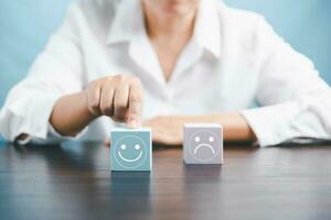 Woman hand holding happy face smile face icon on cube blue object. Customer experience and service with satisfaction concept. positive thinking, mental health assessment, world mental health day. photo
