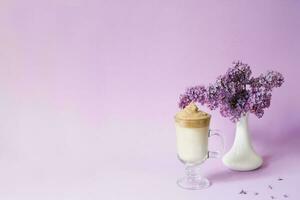 Beautiful dalgona drink a frothy coffee in a transparent mug and flowers lilac to purple Il purple background. Next to the mug is a branch of lilac flowers in a white vase. Copy space photo